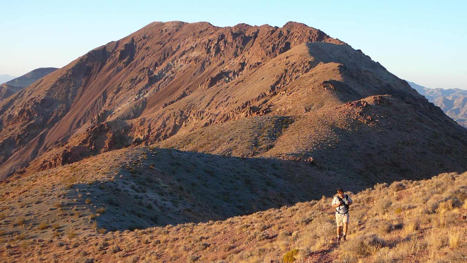 A hiker walks toward the camera on a wide undulating desert ridge covered with small shrubs.