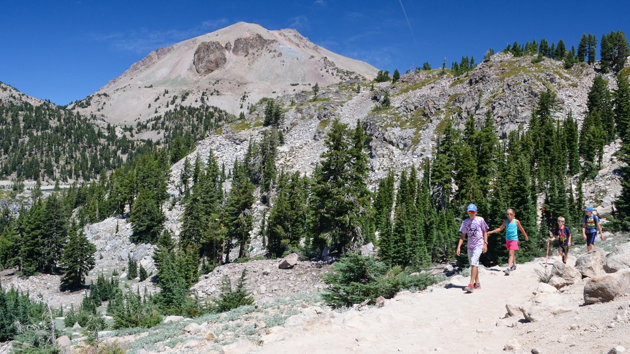 Four young kids hiking on a trail backed by a large volcanic peak and rocky, tree-lined slopes.