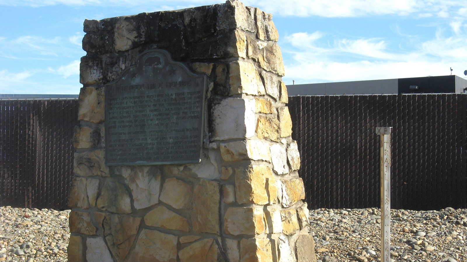 A large stone monument with tapering sides and a plaque on the front stands in a graveled strip.