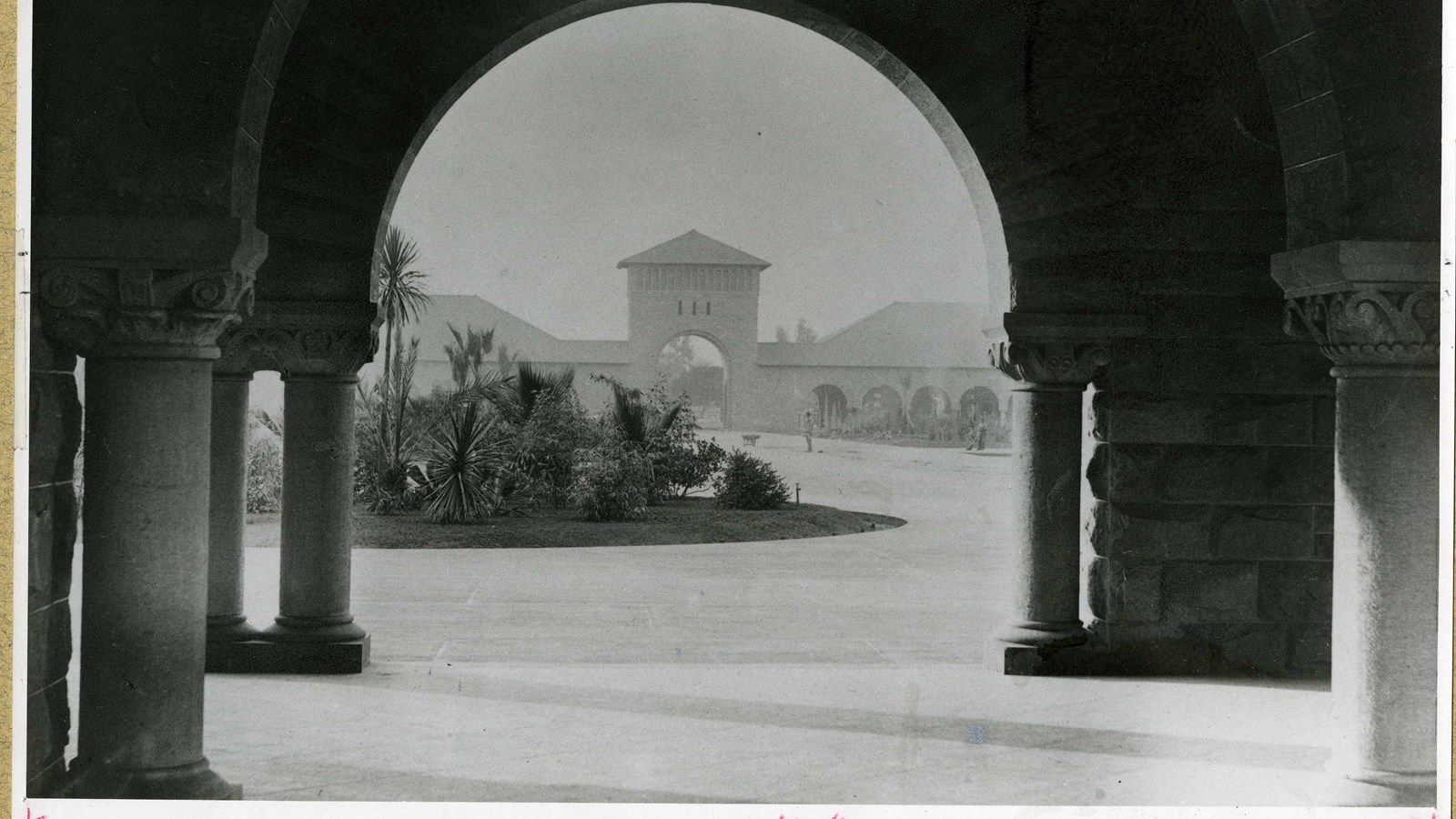 Black and white of stone arch looking across dirt path with grass circle of shrubs