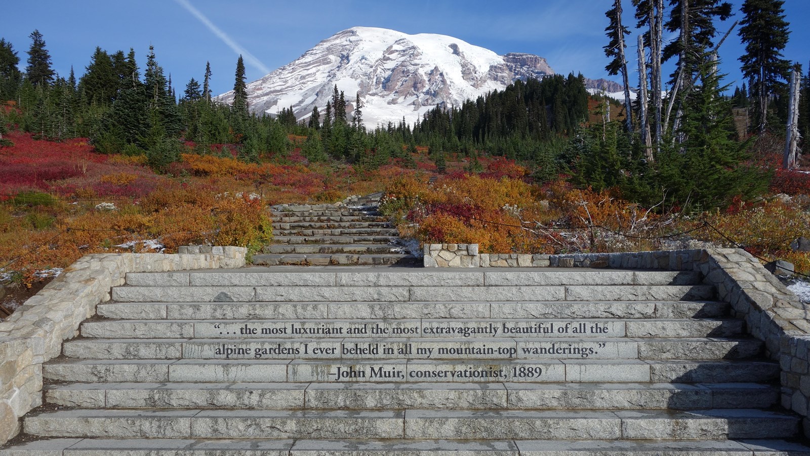 Stone steps lead towards the glaciated peak of Mount Rainier framed by red-orange fall foliage.