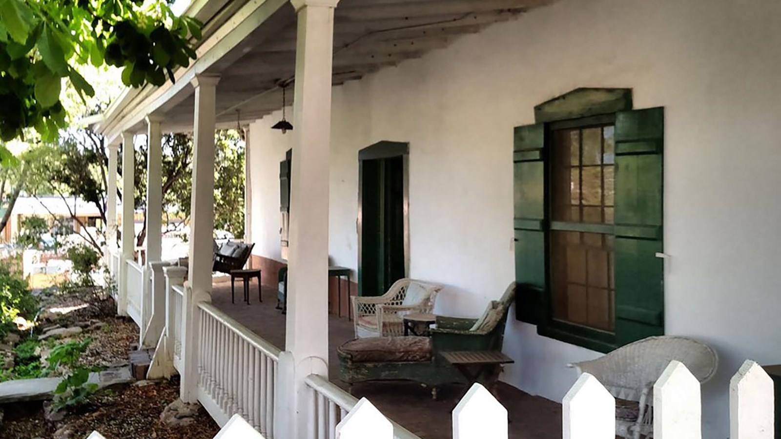 The covered porch of a white stucco house with a white railing and picket fence.