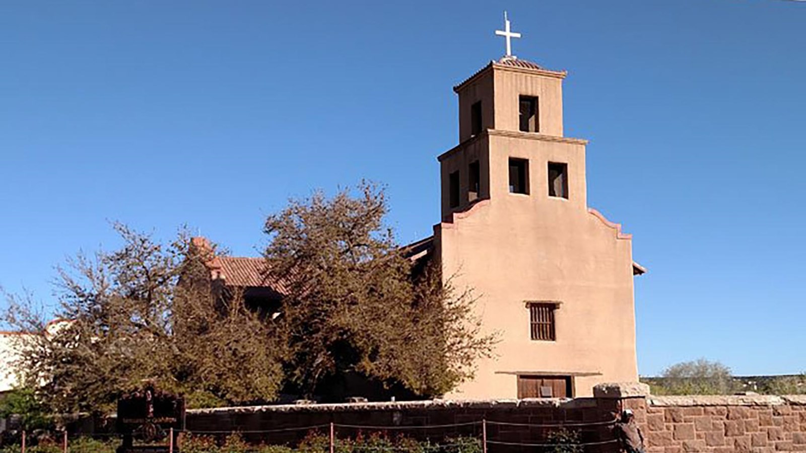 A large, single bell-towered, adobe chapel with a large stone wall surrounding it.