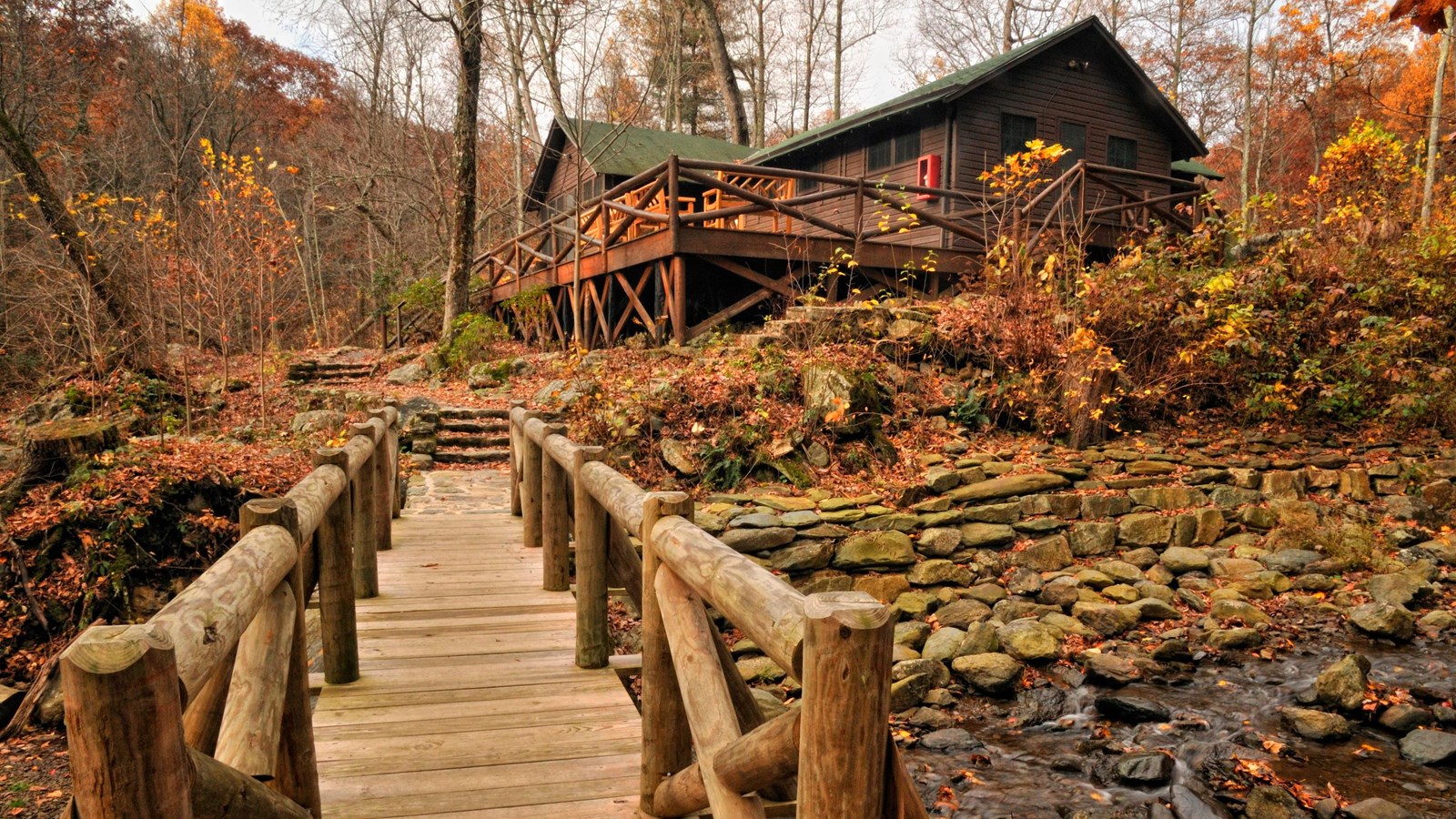 A wooden bridge leads across a mountain stream to a brown cabin in the woods.