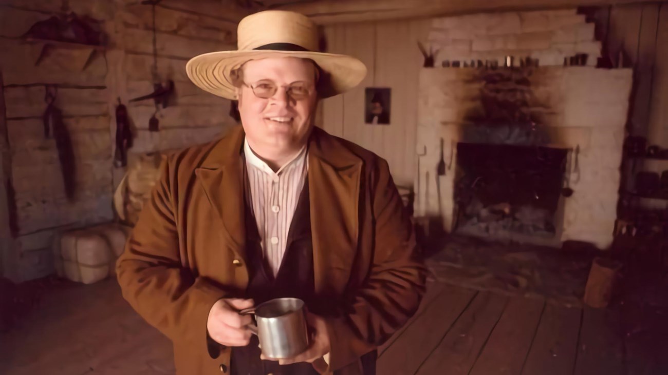 Man in straw hat brown wool coat, vest and striped shirt holds a tin cup