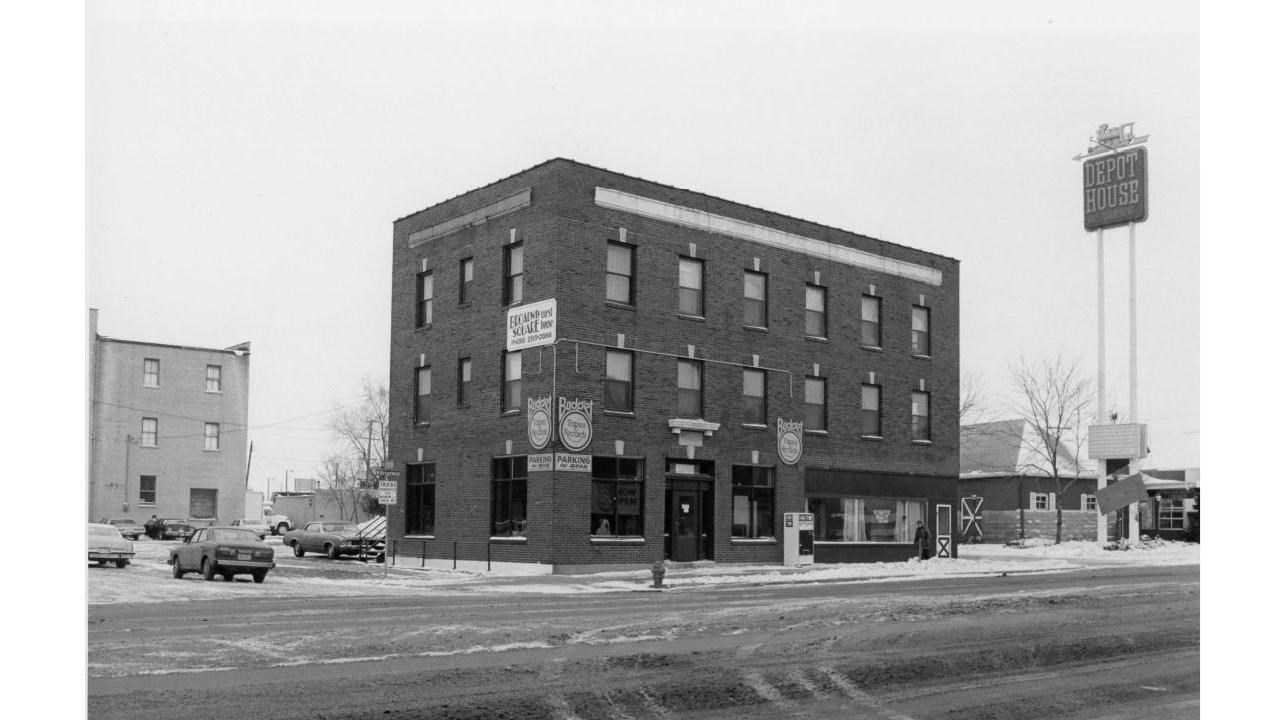 three story brick building on the side of a road with a parking lot and cars 