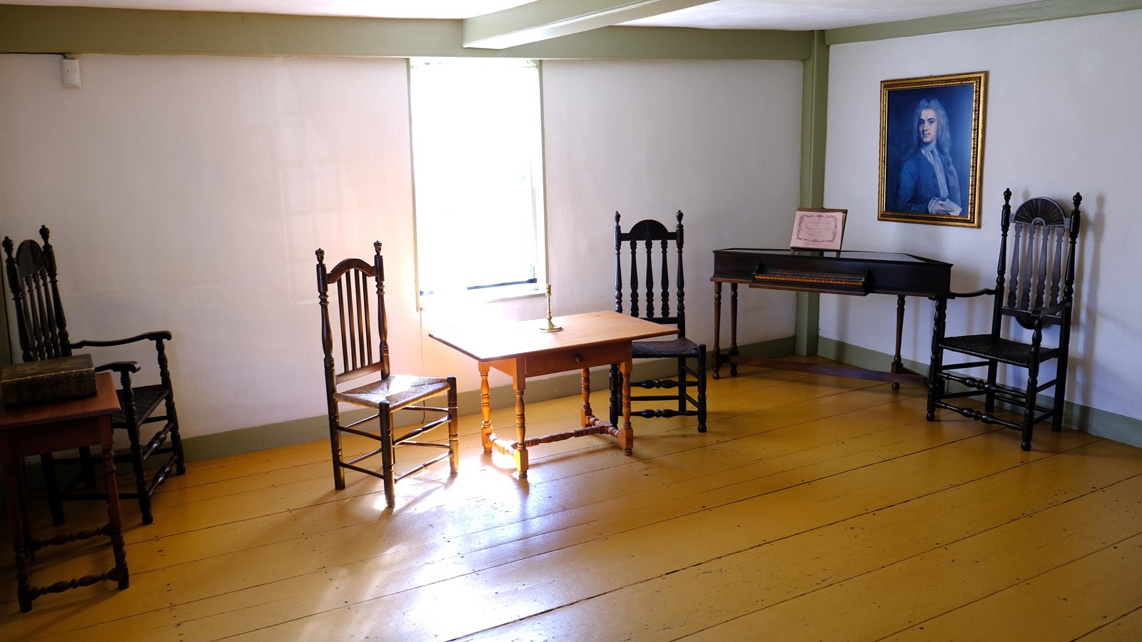 A room with white walls and four wooden chairs, tables, harpsichord, and a portrait on the wall.
