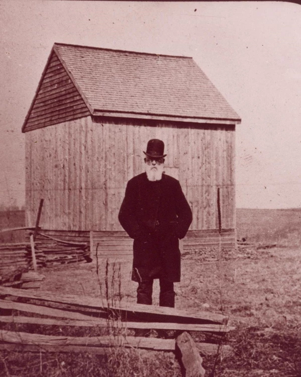 Older man with a white beard and hat with a wooden barn in the background.