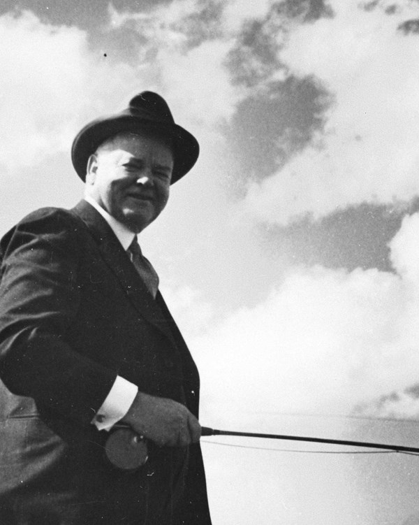 A black and white photo depicts a smiling middle aged man in a hat outdoors.