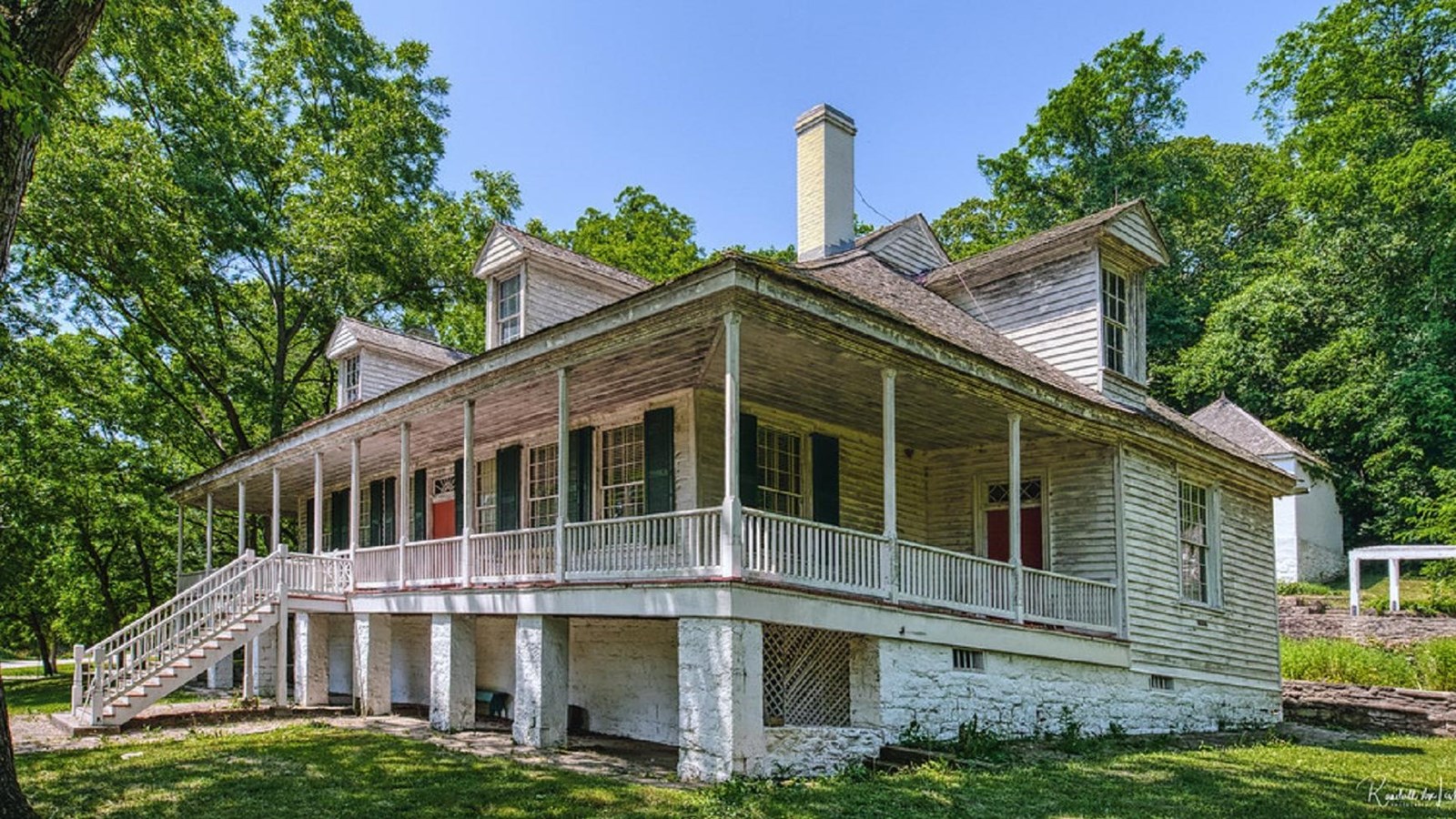 A two story colonial house with a wrap-around porch, dormers, and a chimeny admit trees and grass
