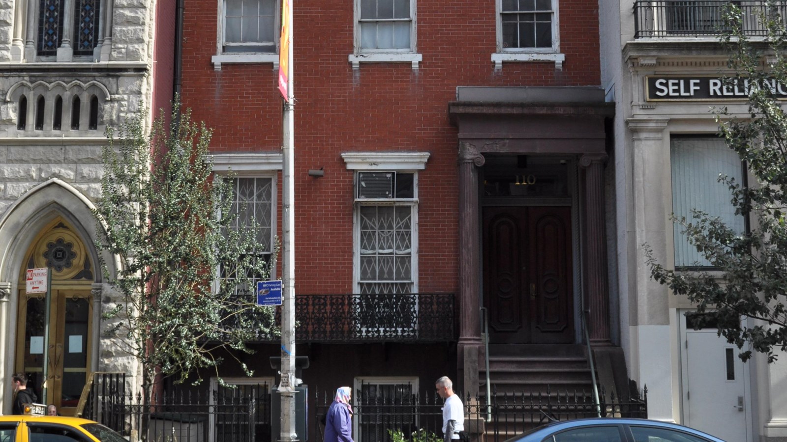 Three story red brick building with 3 windows per level in between a church and another building.
