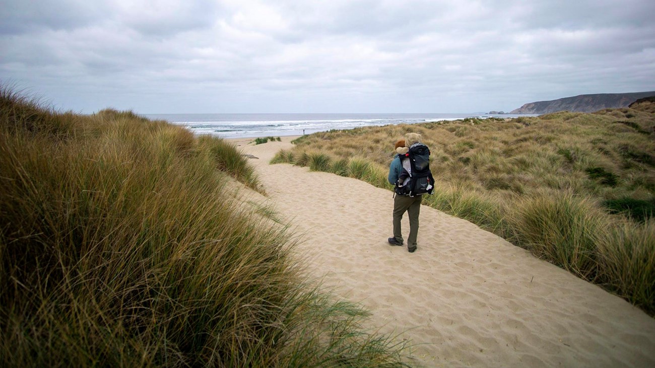 A photograph of a man carries a young child on his back while walking along a path over a sand dune.