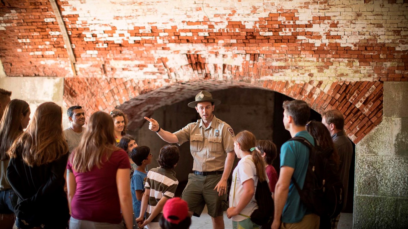 A uniformed ranger speaks to a crowd of mixed ages in an old brick hallway. 