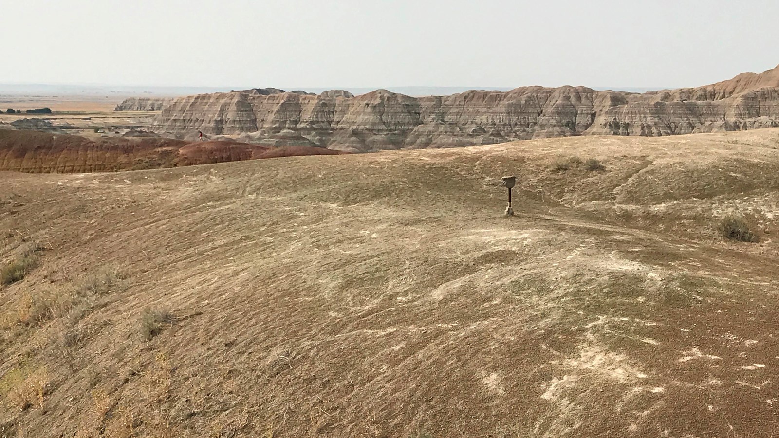 Two metal poles extend out of a rolling hill under hazy sky with badlands formations in horizon.