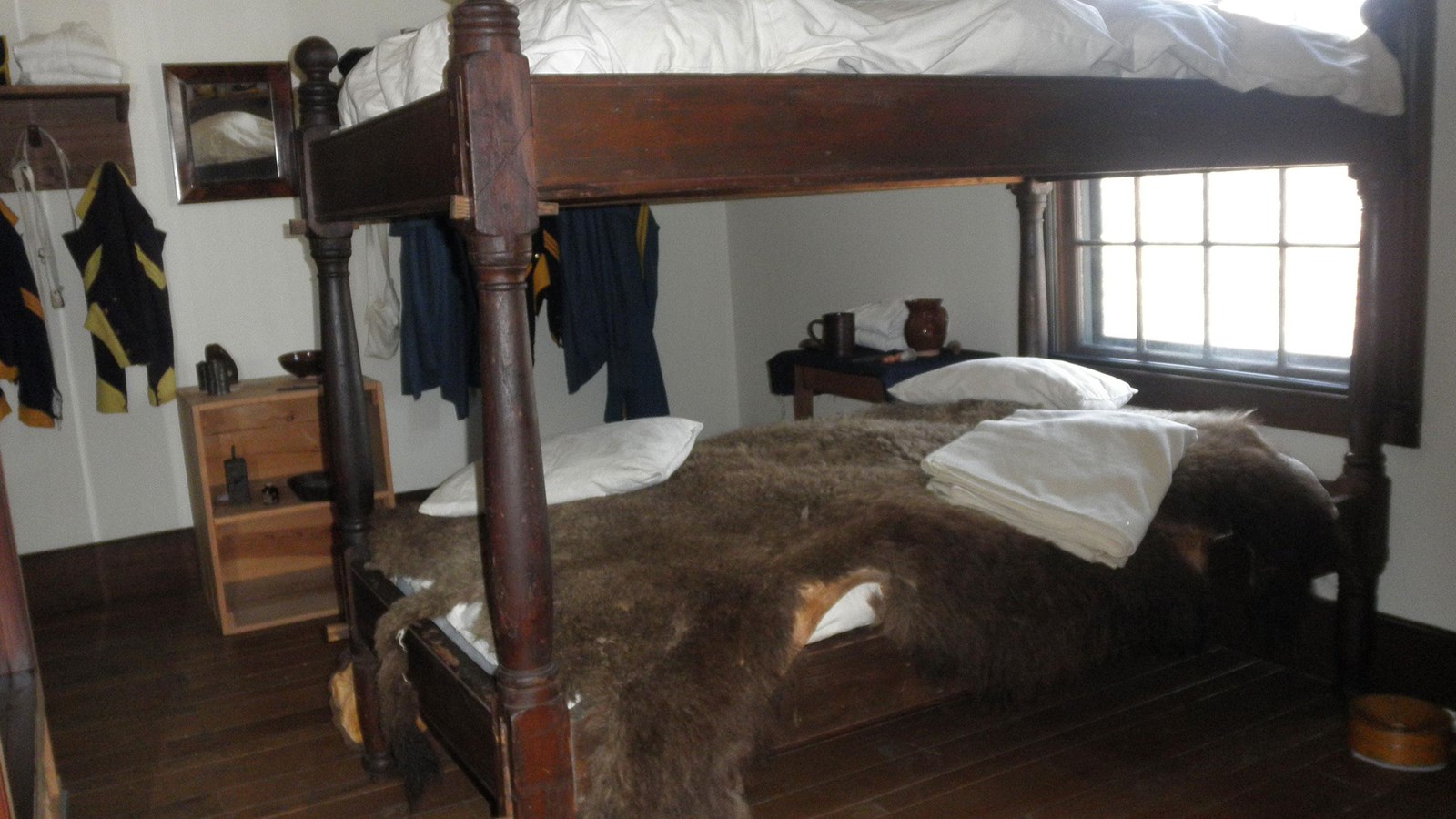 Wooden bunk bed with buffalo robe.  Uniforms hanging on pegs on wall.