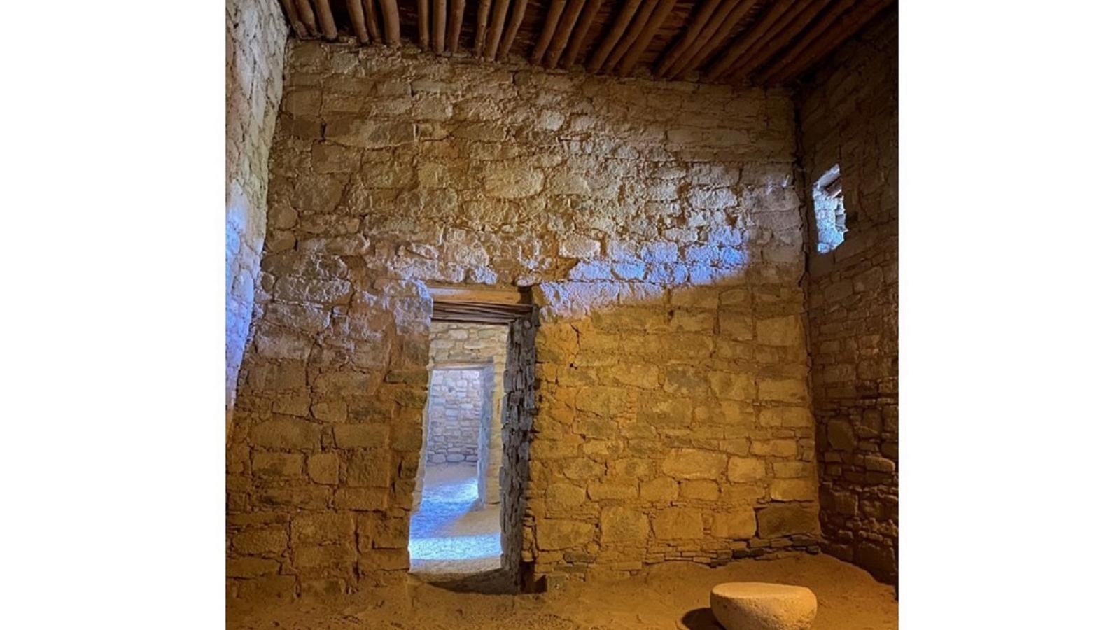 The interior of a room with tall sandstone walls, a grinding stone on the floor, & a wooden ceilin