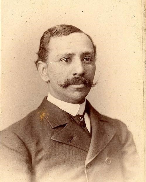 Sepia-tinted photograph of a man from the chest up, wearing a suit and with a prominent mustache.