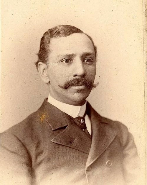 Sepia-tinted photograph of a man from the chest up, wearing a suit and with a prominent mustache.