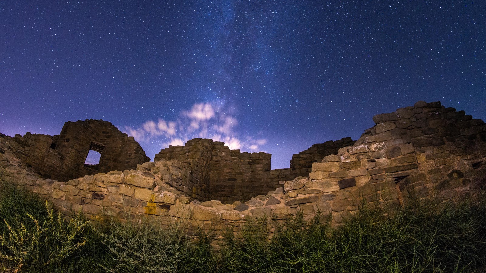 A purple night sky sitting above a sandstone stacked wall with clouds and stars shining bright.