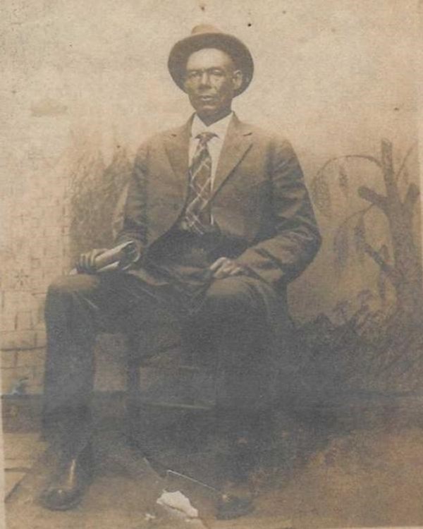 Man sits wearing a suit and tie and a derby hat.