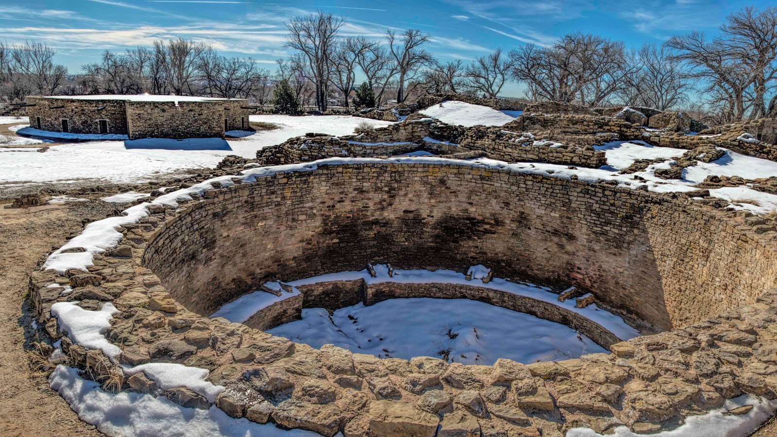 A circular sandstone kiva and other sandstone remnants of the structure in the background with snow.