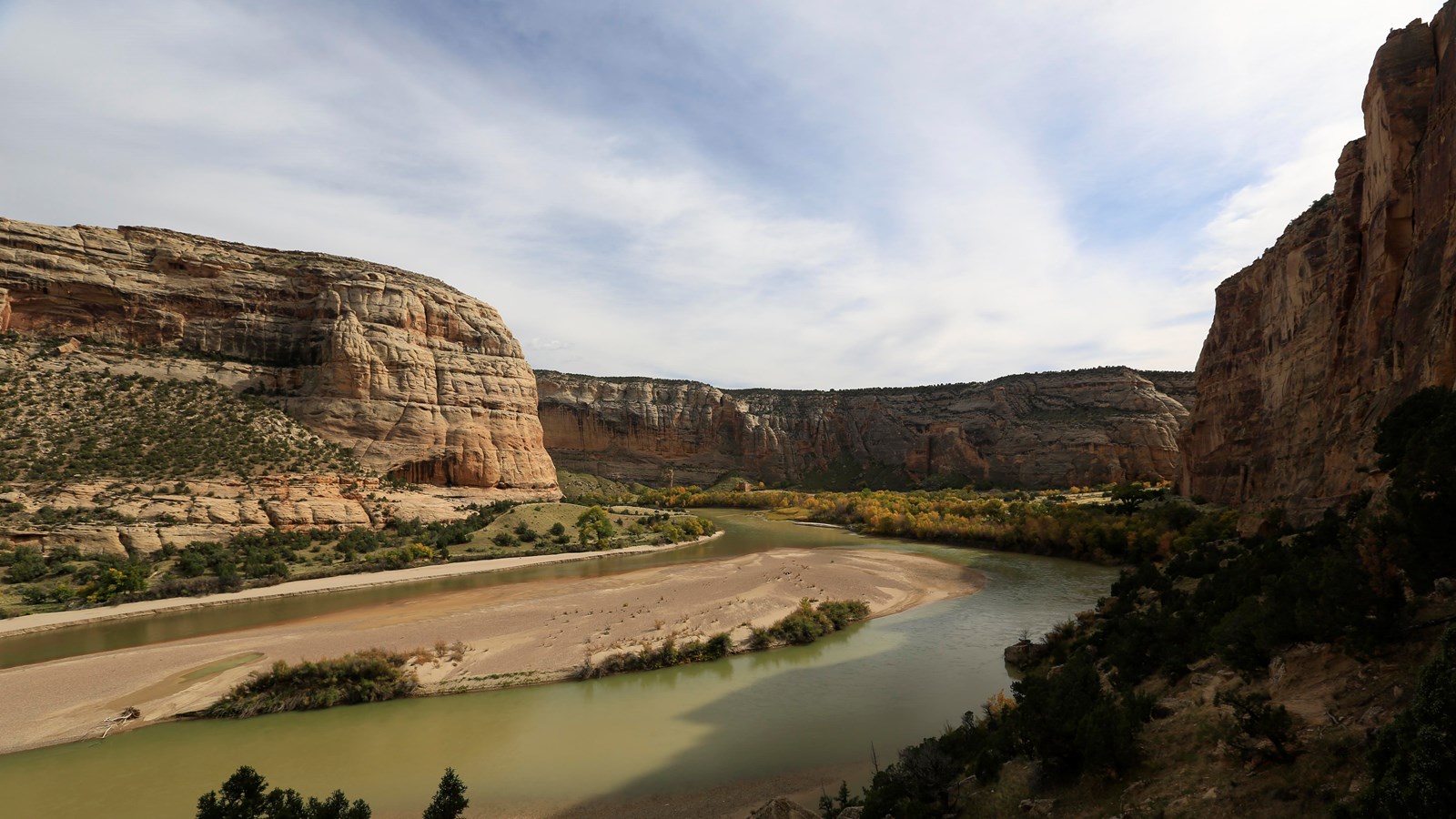 A sandstone canyon with a wide river containing long sand bars, flowing through.