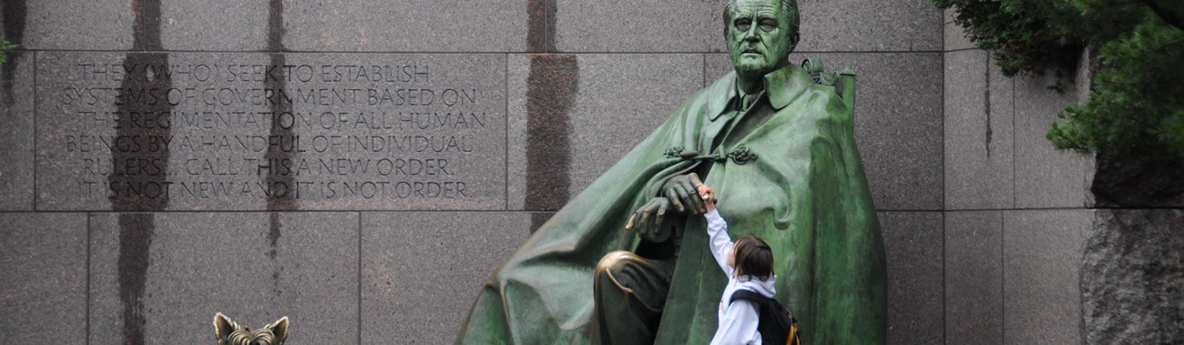 A child reaches up to touch a large bronze statue of Franklin Roosevelt.