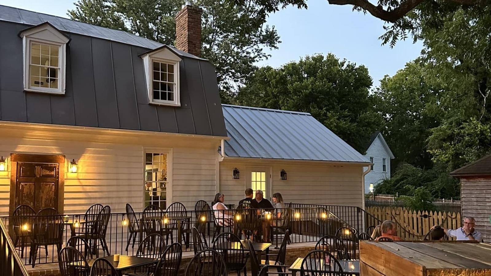 Outdoor dining patio with tables, chairs, and string lights next to a white house.