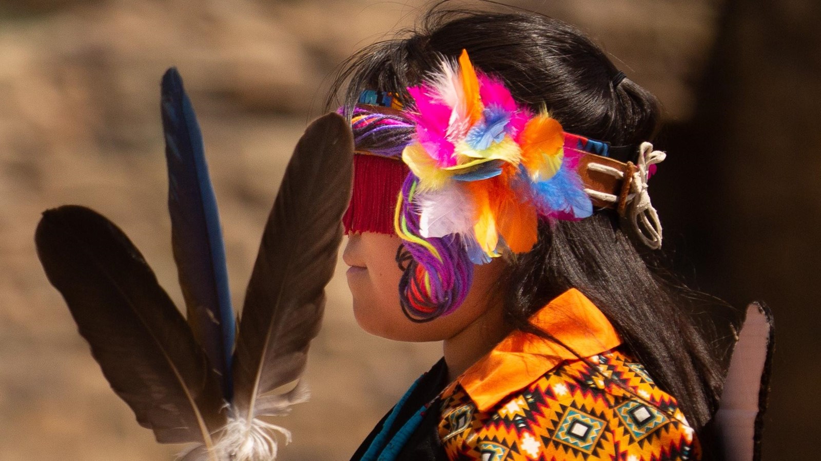 A young girl dressed in traditional dance clothing holding feathers & wearing a covering on her eyes