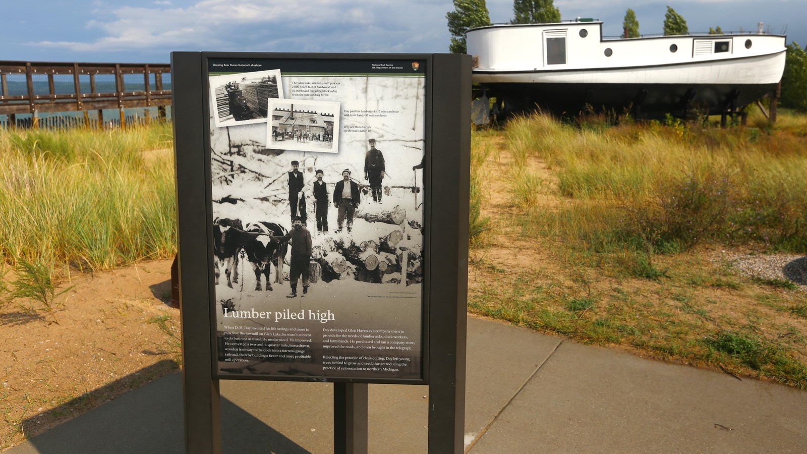Interpretive panel stands in front of swath of dune grass and the tug Aloha