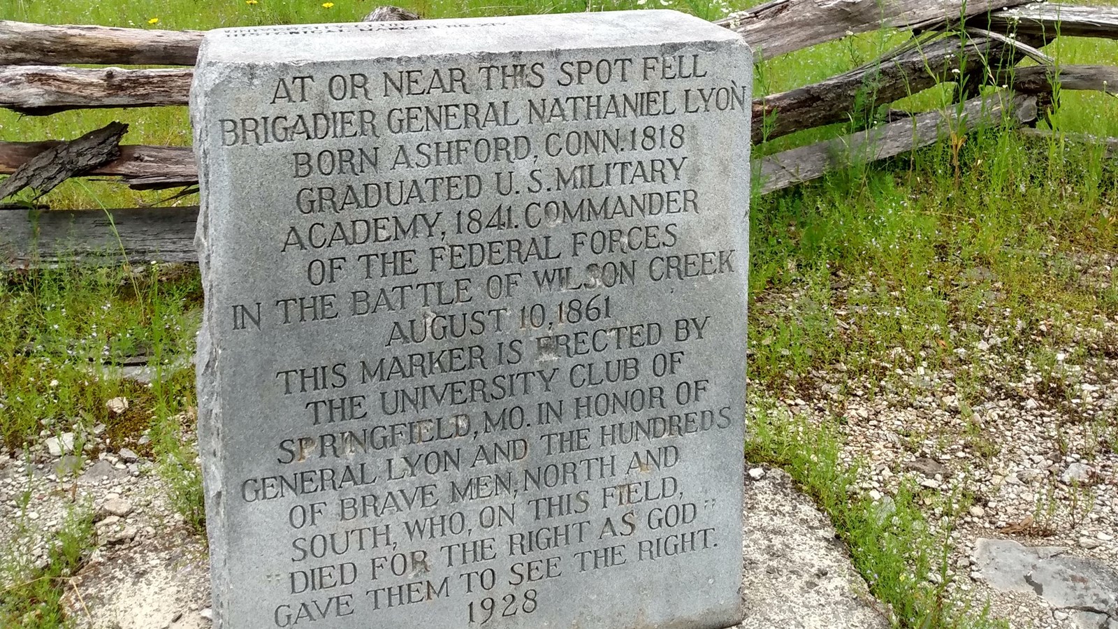 A plain stone marker with engraving sits in front of a split-rail fence in an open area