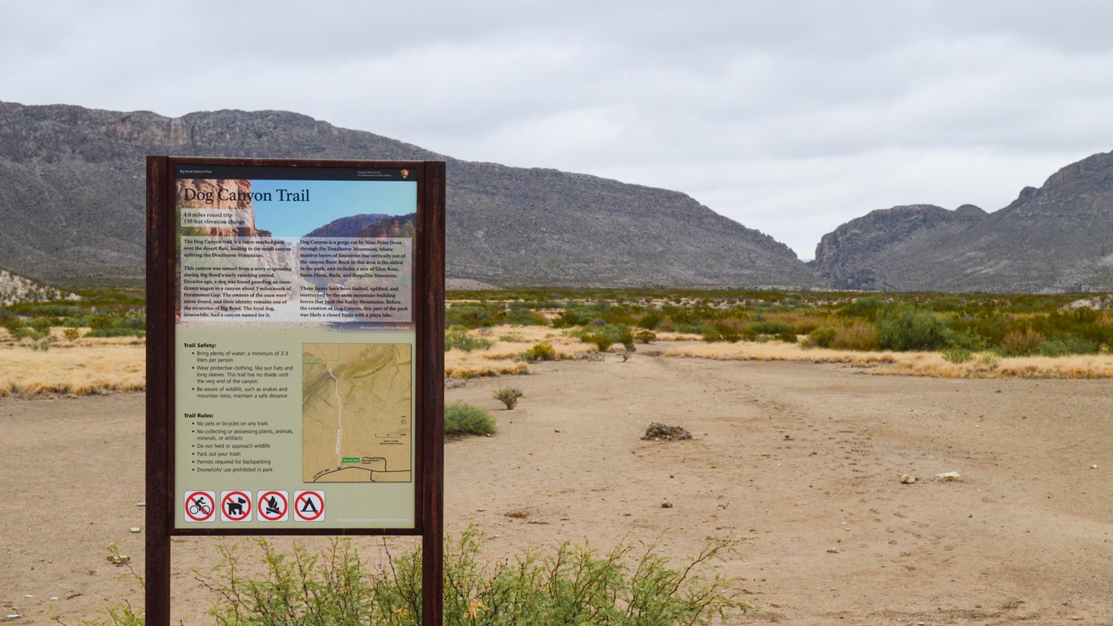 A metal sign with trail information is in front of a flat desert trail that leads to a canyon.