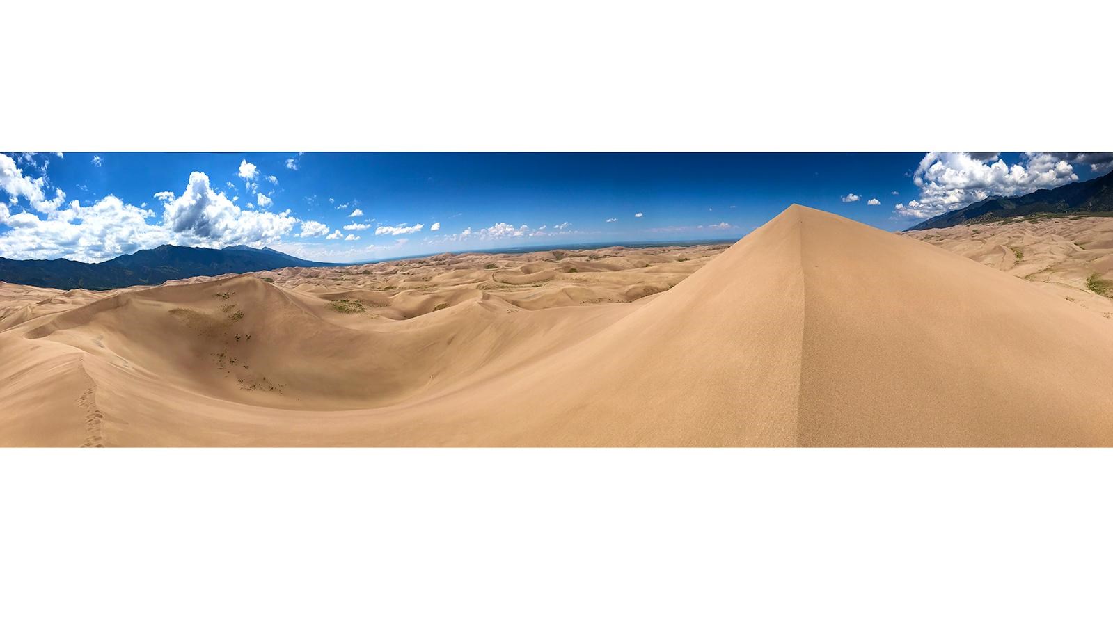 A panoramic view of dunes, with mountains in the distance and blue sky above. 