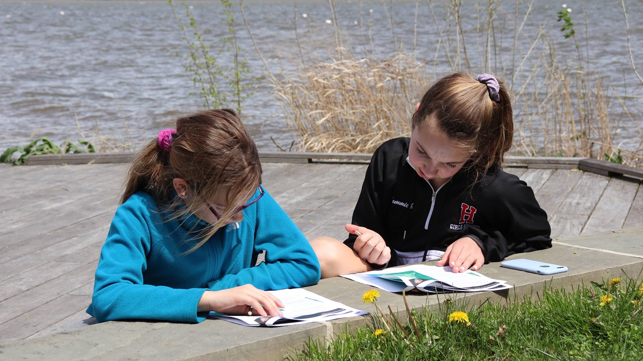 Two children working on a booklet with a flowing river in the background