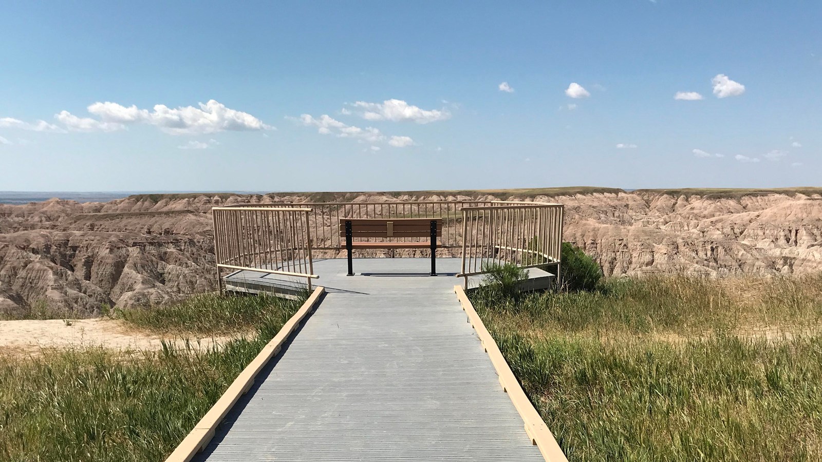 A single boardwalk extends out to a bench which sits against a vast landscape under blue sky.
