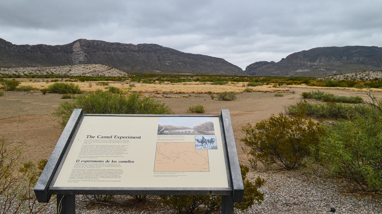 A metal sign with historic information sits in the foreground of a desert and mountain landscape.
