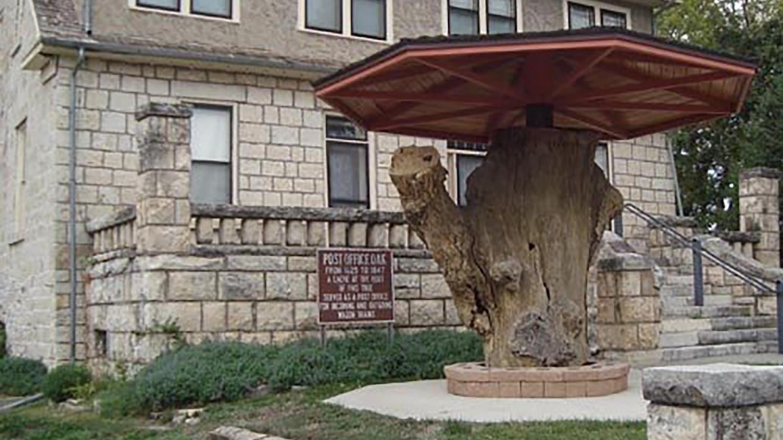 The stump of a large tree sits under a piece of wood in front of a building.