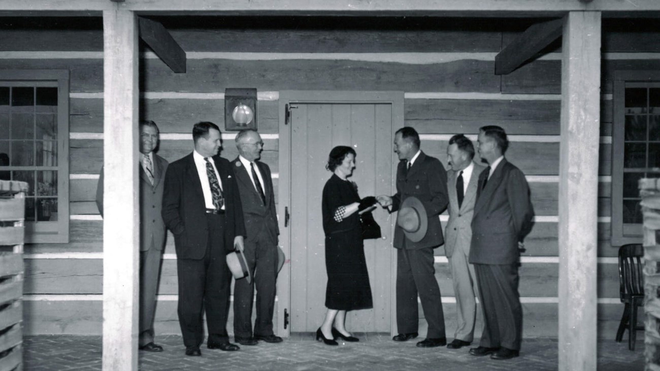 Woman in a black dress stands in front of a cabin door, three men in suits stand on each side of her