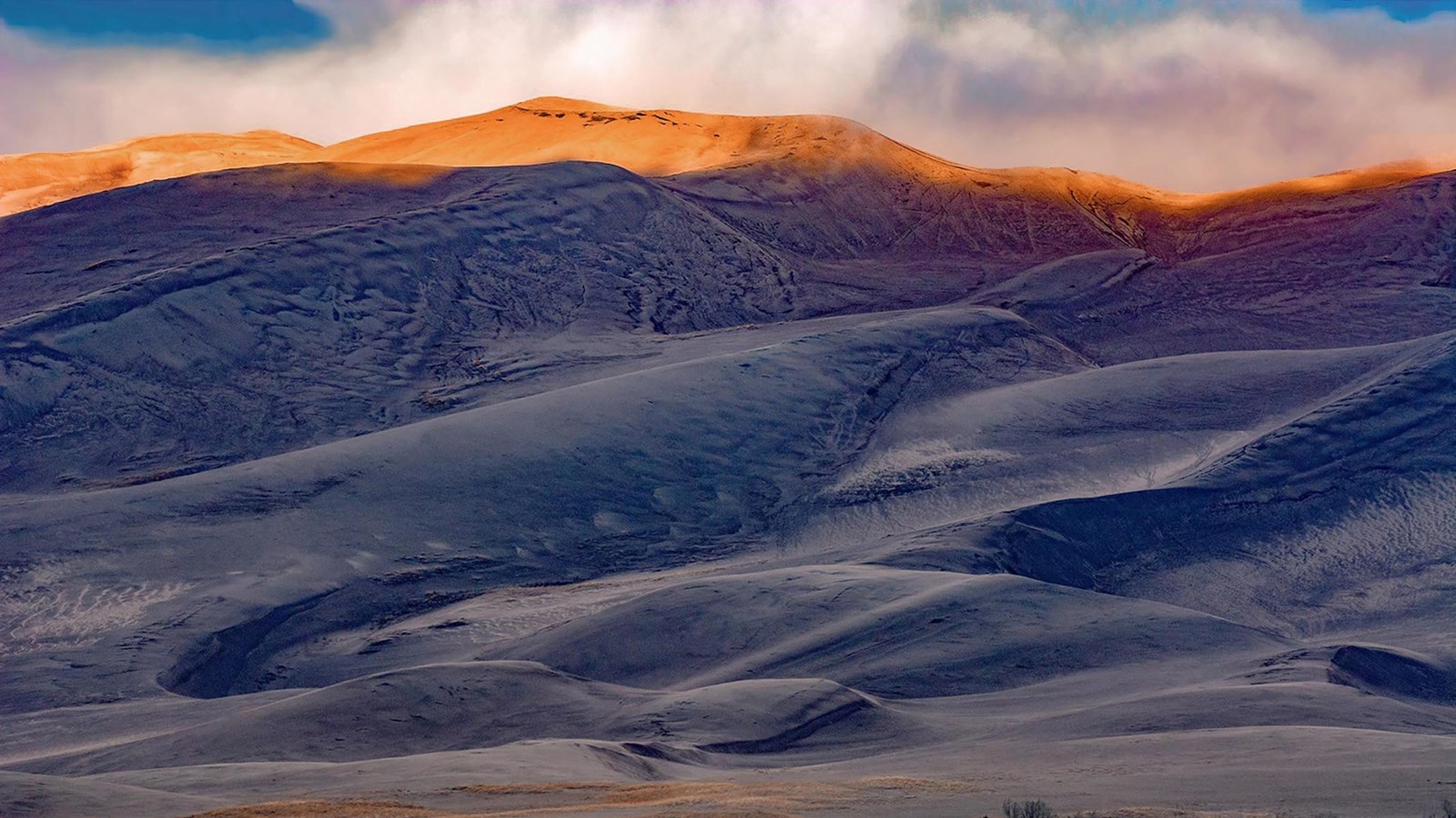 The top of a tall dune is illuminated by the rising sun, as clouds swirl in blue sky