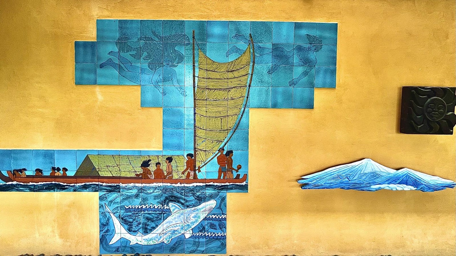 Mural showing polynesian voyagers, white shark, snow capped mountains, and a wooden carving of sun.