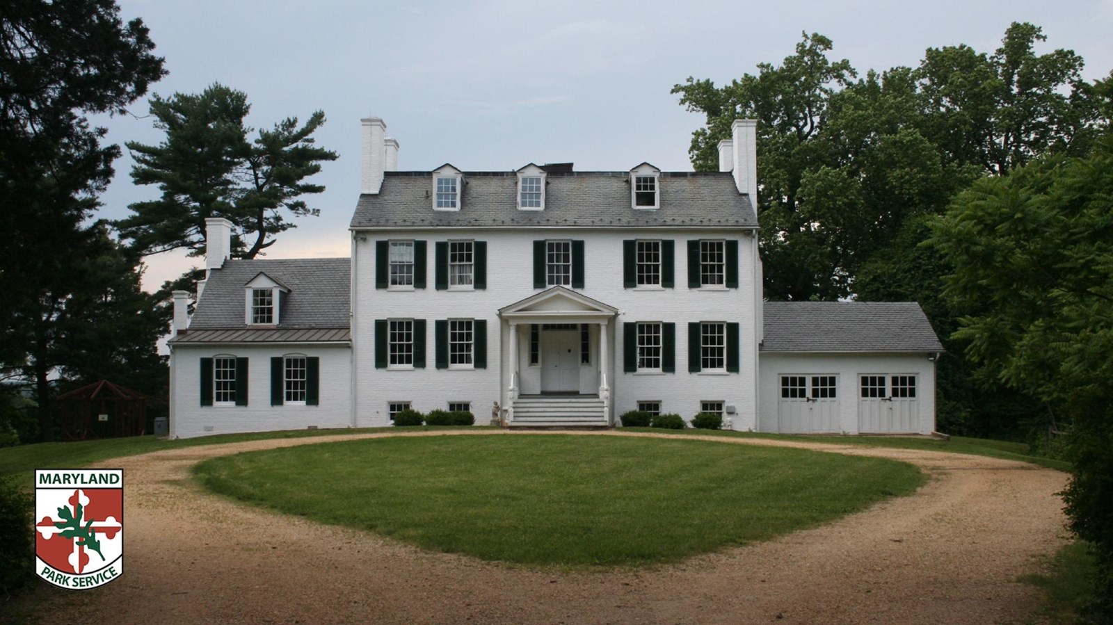 White historic house with green shutters, surrounded by trees and a circular gravel driveway.
