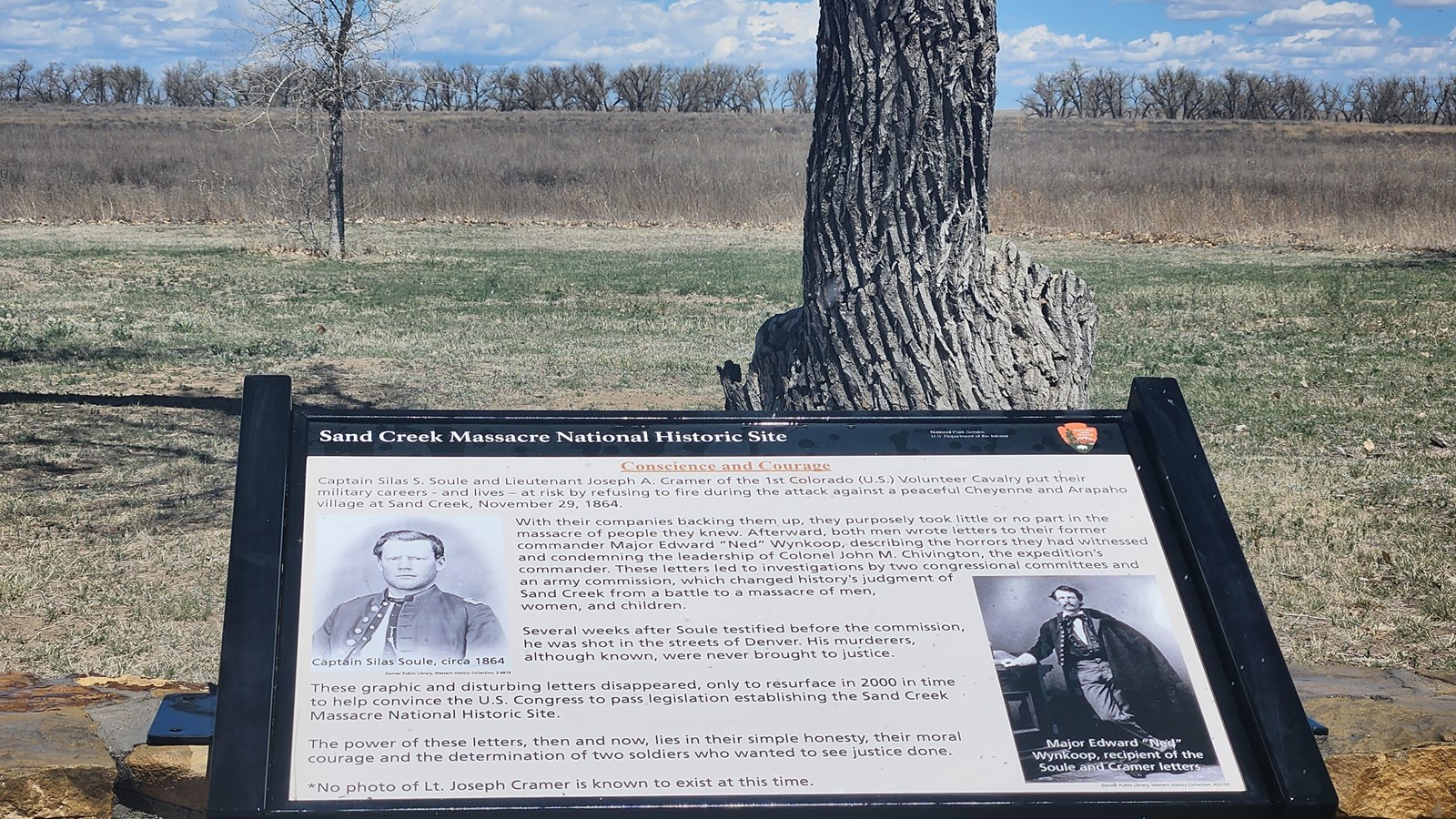 A sign in the foreground with open prairie beyond it, and a line of trees in the far distance.