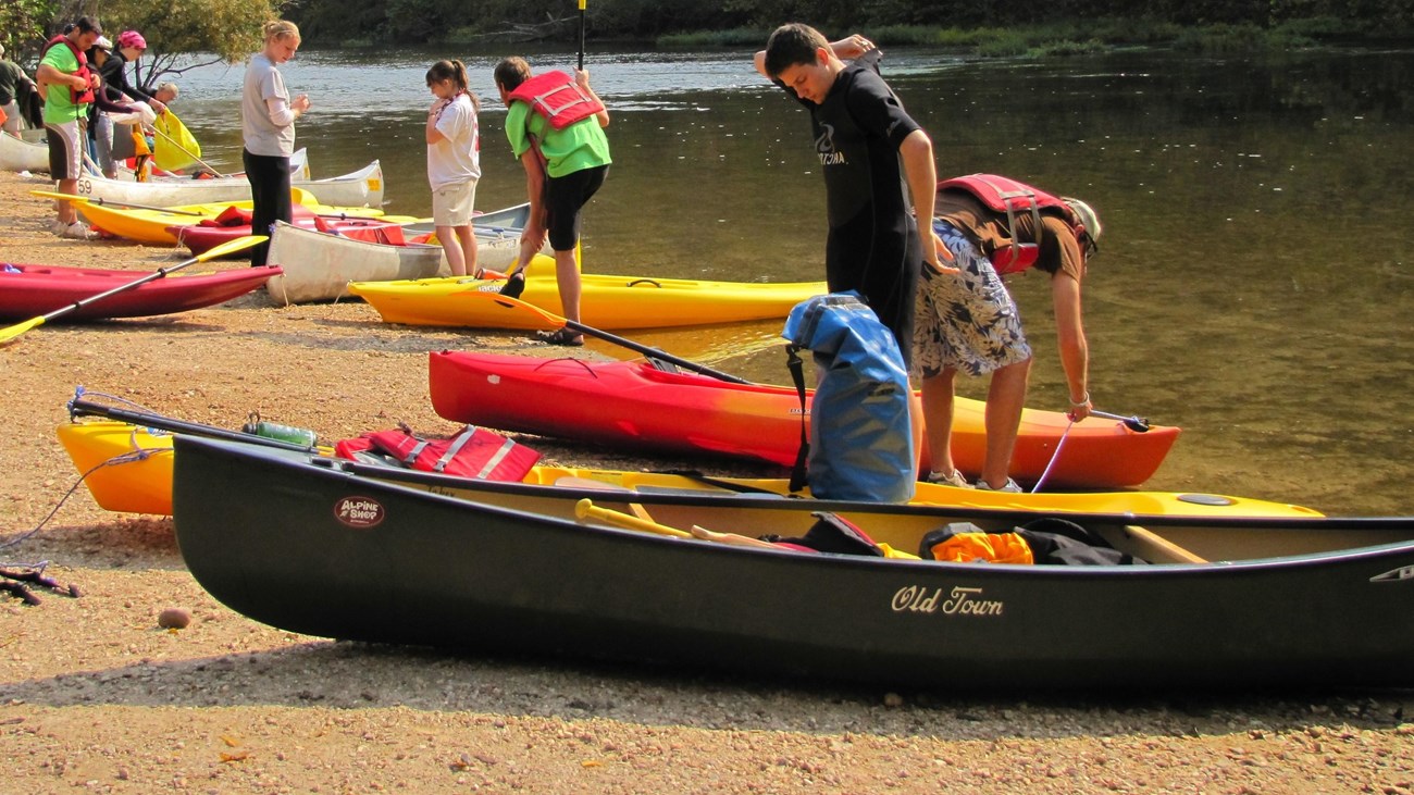 a group of young people on shore next to river with canoes and kayaks