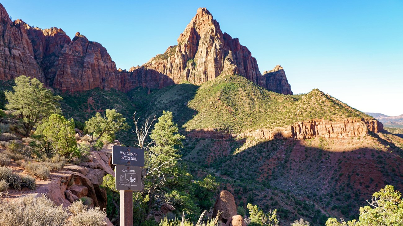 View from a sandy overlook of a triangular sandstone mountain with green foliage on the foothills.