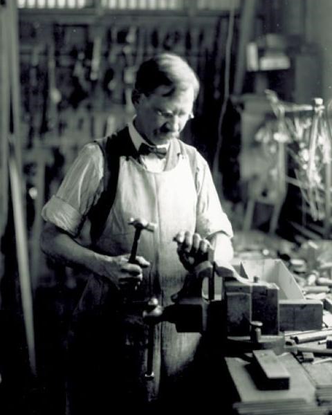 Charlie Taylor, a man of medium-build wearing a white shirt and leather apron, works at a workbench