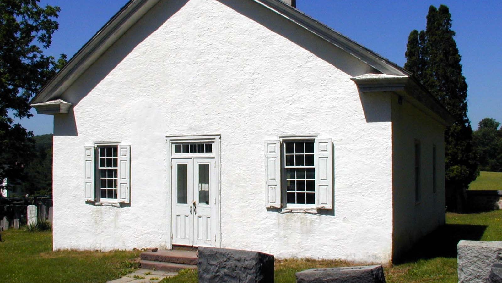 A white building with two windows and a double door. The building is surrounded by grave markers. 
