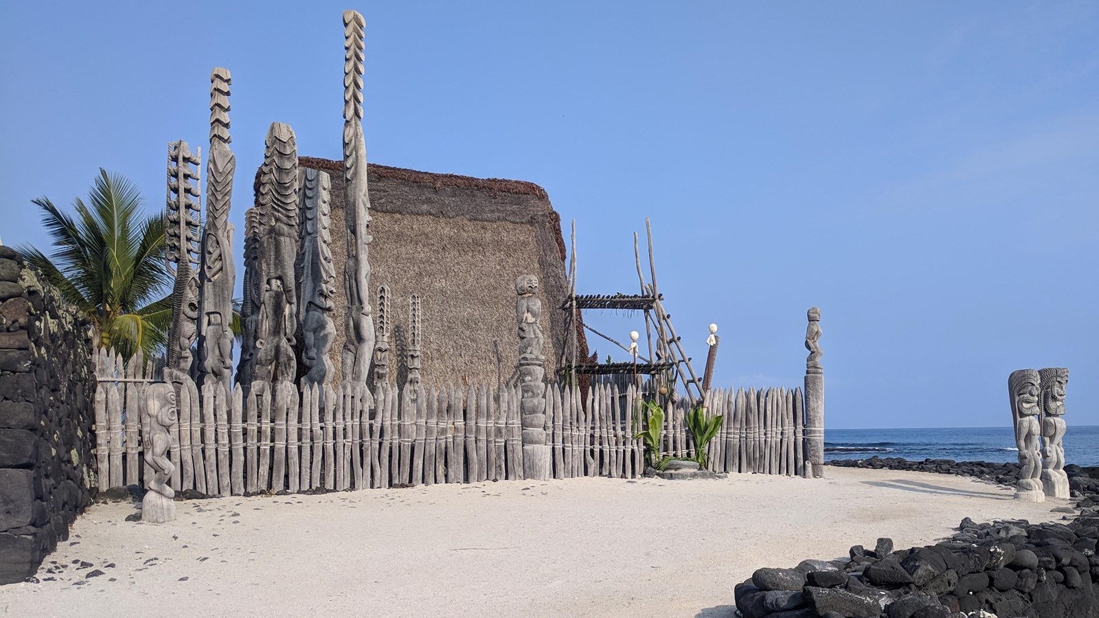 The thatched royal mausoleum sit within a wooden fence line with intricately carved kiʻi images