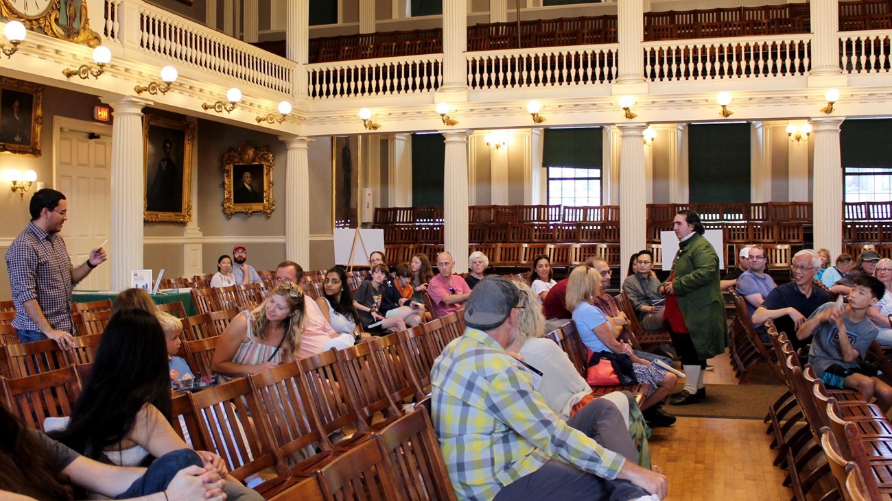 People sitting in chairs in a great hall with a Ranger dressed in period clothing.