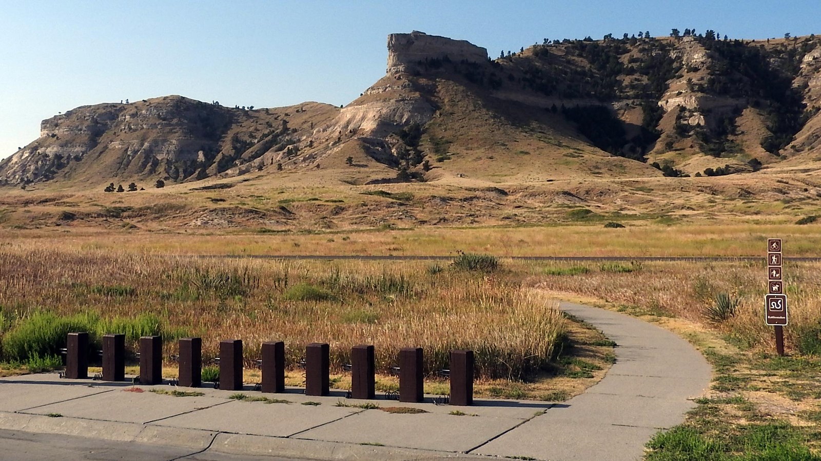 A bicycle rack is seen to the right of a concrete trail with rock formations in the background