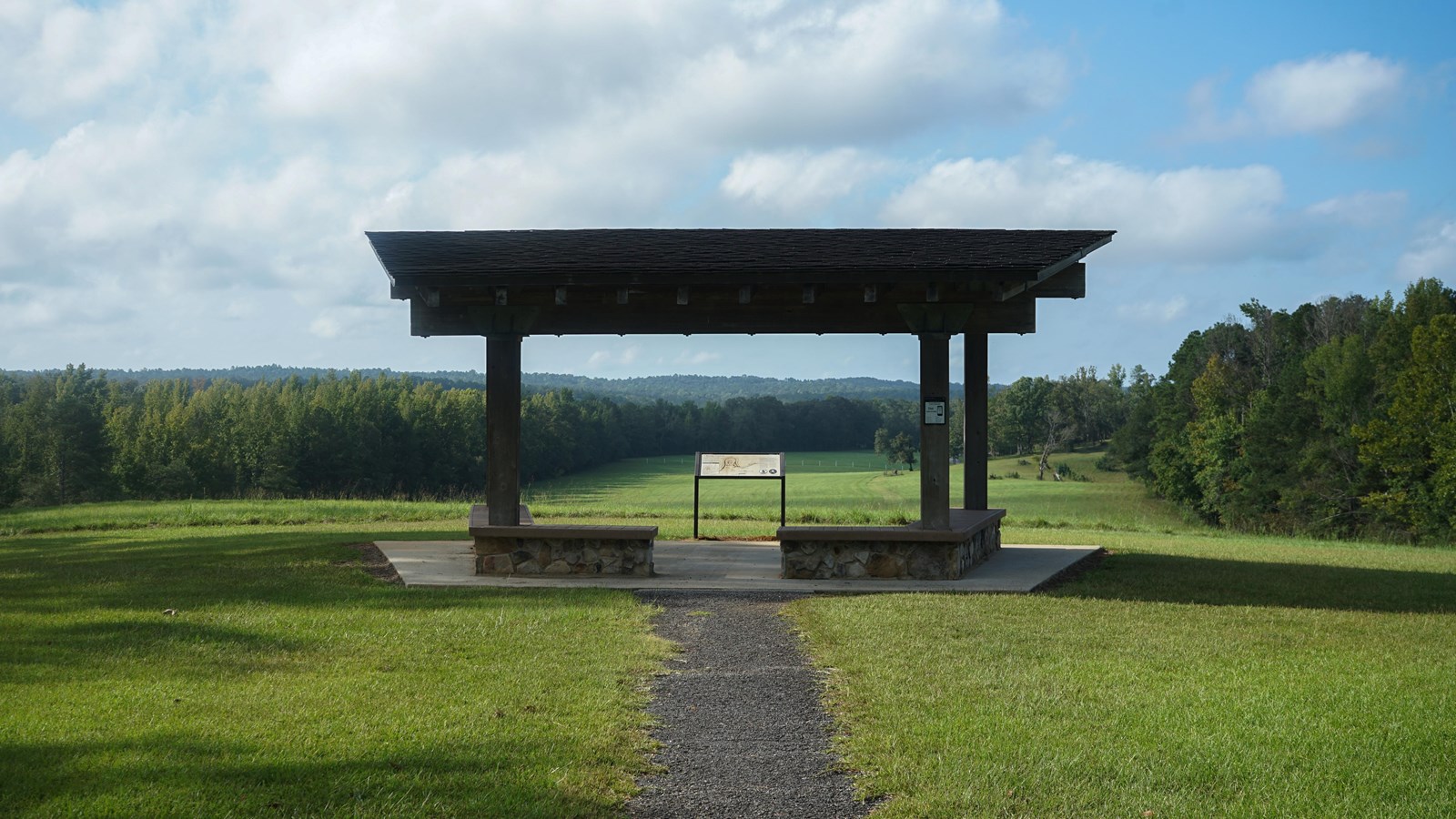Image displays an informational wayside surrounded overlooking a grassy hill and forested area.